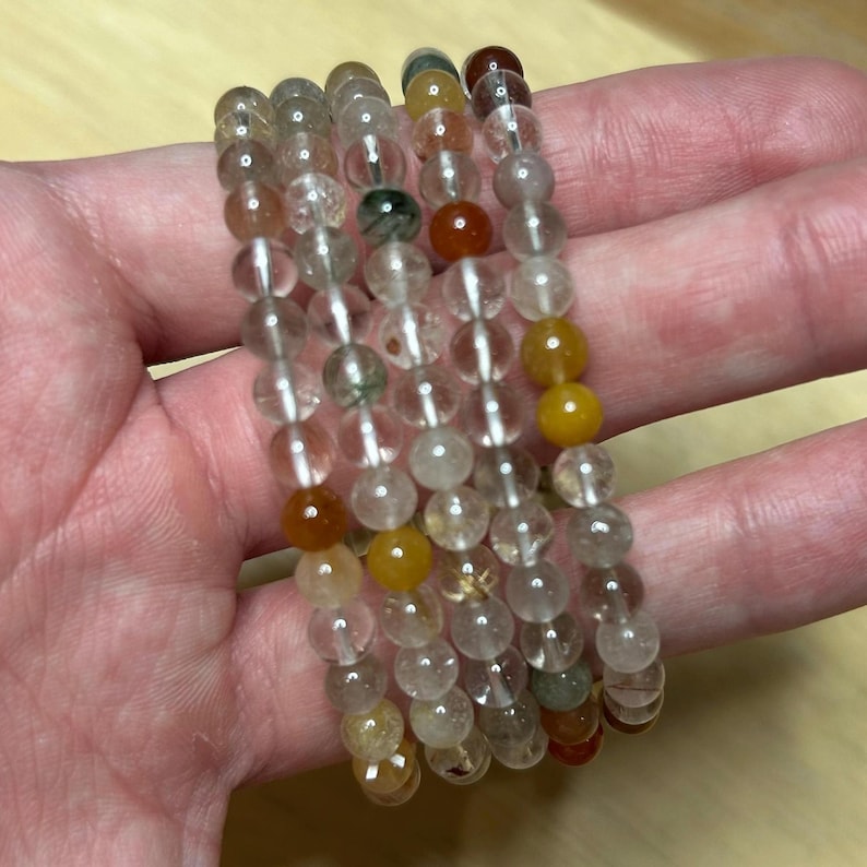 May include: Five beaded bracelets with clear, yellow, brown, and green beads. The bracelets are made of small, round beads strung together. The bracelets are held in a hand.