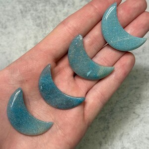 May include: Four blue-green crescent moon-shaped stones. The polished stones have a speckled appearance and a gradient of color, transitioning from a darker blue-green to a lighter, almost white hue. The stones are held in a hand.