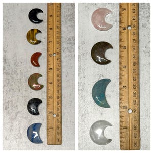 May include: Assortment of polished gemstone crescent moons in various colors, including black, tiger's eye, red, and blue. A wooden ruler with inch measurements is placed next to the gemstones. The moons are approximately 1 inch in length.