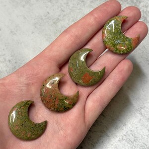 May include: Four polished unakite crescent moon stones. The stones have a mix of green, red, and orange hues. The crescent moon shape is smooth and curved, with a glossy finish. The stones are held in a hand.