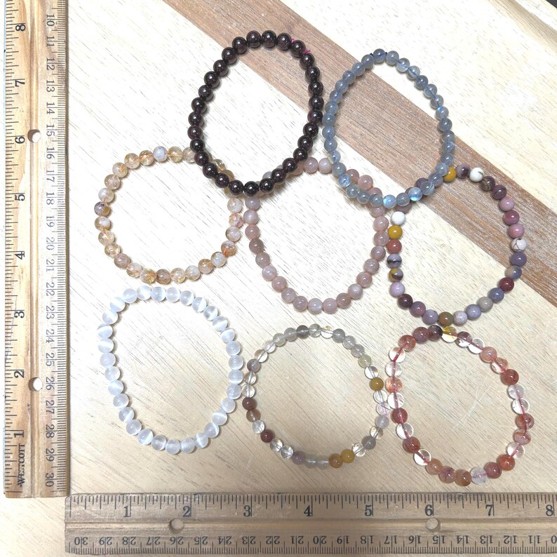 May include: A collection of eight beaded bracelets in various colors, including brown, pink, and clear. The bracelets are arranged on a wooden surface with a ruler for scale. Each bracelet appears to be made of small, round beads strung together.