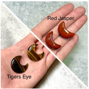 May include: Three crescent moon-shaped gemstones: two Tiger's Eye and two Red Jasper. The Tiger's Eye stones have brown and gold stripes. The Red Jasper stones are a deep red color. The text "Tigers Eye" and "Red Jasper" are in the image.
