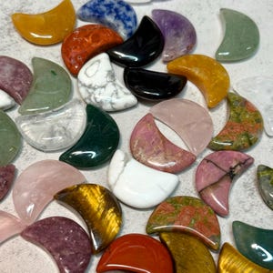 May include: A collection of crescent-shaped gemstones in various colors and patterns. The stones include shades of yellow, orange, green, pink, purple, white, and black. The gemstones are arranged on a light gray surface.