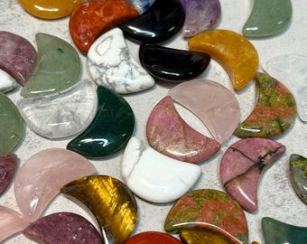 May include: A collection of crescent-shaped gemstones in various colors and patterns. The stones include shades of yellow, orange, green, pink, purple, white, and black. The gemstones are arranged on a light gray surface.