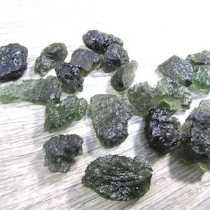 May include: A collection of rough, dark green moldavite gemstones. The stones have irregular shapes and a textured surface, with varying shades of green and hints of translucence. The gemstones are scattered across a light-colored surface.