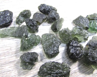 May include: A collection of rough, dark green moldavite gemstones. The stones have irregular shapes and a textured surface, with varying shades of green and hints of translucence. The gemstones are scattered across a light-colored surface.