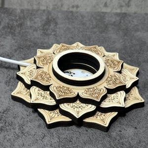 May include: A wooden, laser-cut lotus flower-shaped wireless charger. The charger has a central circular opening and intricate floral patterns etched into the wood. A white charging cable extends from the base. The charger is approximately 5 inches in diameter.