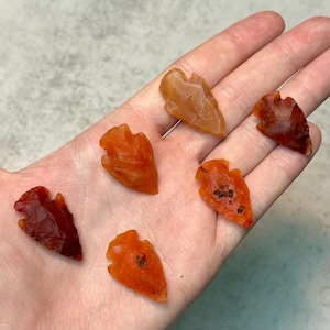 Puede incluir: Seis pequeñas puntas de flecha de color naranja y rojo. Las puntas de flecha están hechas de una piedra translúcida, con variaciones de color y textura. Las puntas de flecha se muestran sobre un fondo claro.
