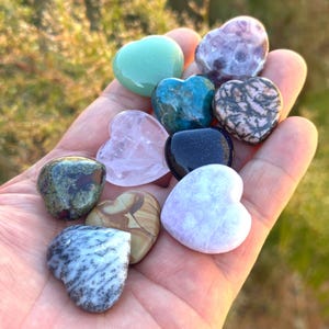 May include: A collection of heart-shaped gemstones in various colors, including green, purple, blue, pink, and black. The stones are held in a hand, showcasing their polished surfaces and unique patterns. The gemstones are a variety of sizes and textures.