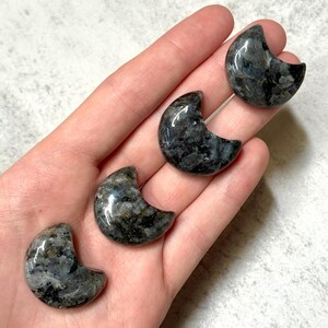 May include: Four polished labradorite crescent moon stones. The stones are a mix of dark gray, black, and iridescent blue-green. The crescent moons are smooth and have a glossy finish. The stones are held in a hand.
