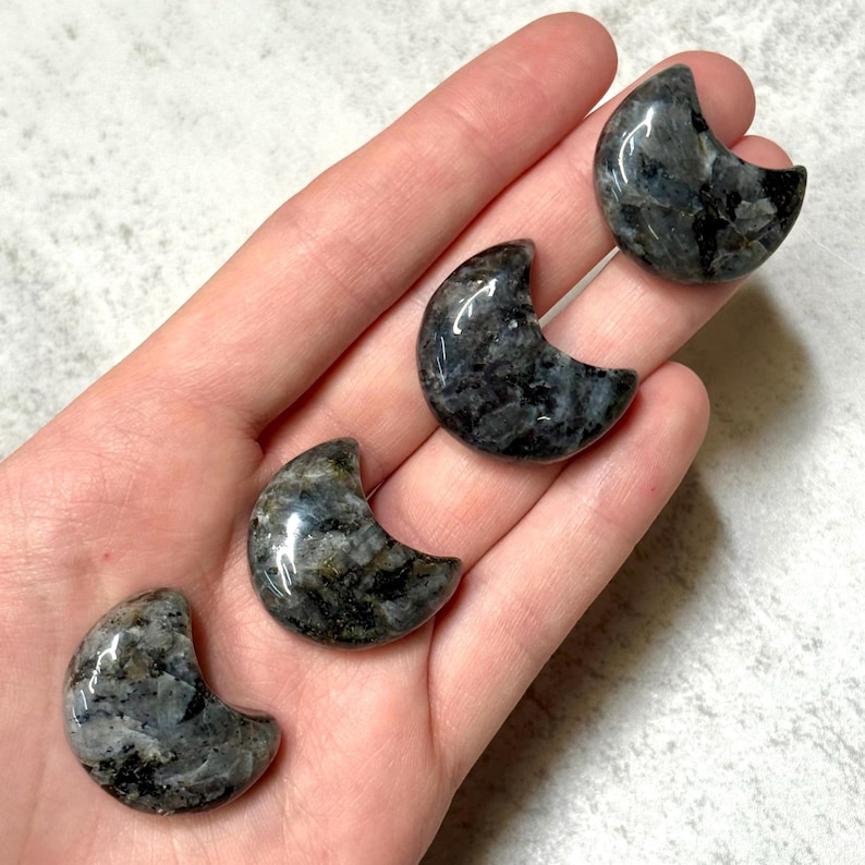 May include: Four polished labradorite crescent moon stones. The stones are a mix of dark gray, black, and iridescent blue-green. The crescent moons are smooth and have a glossy finish. The stones are held in a hand.