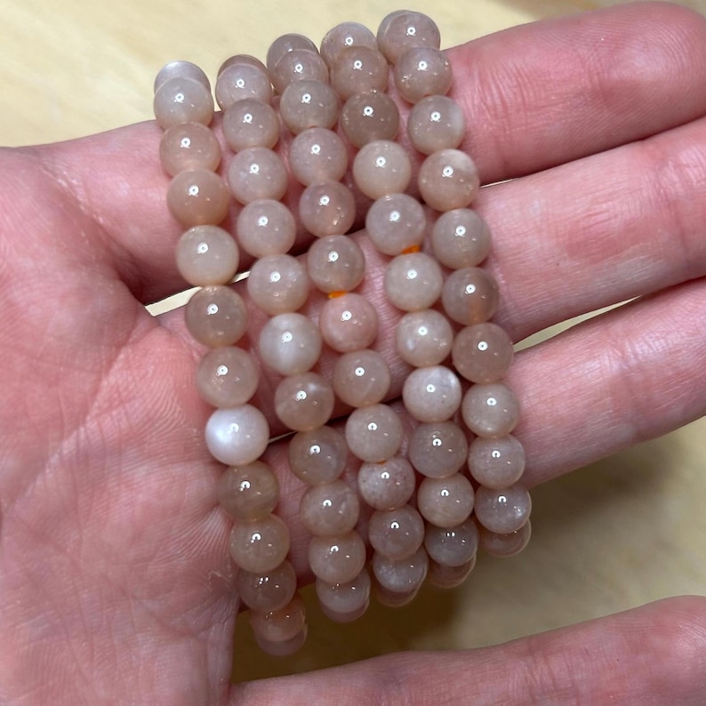 May include: Several strands of peach-colored, round gemstone beads are held in a hand. The beads have a smooth, polished surface and a subtle sheen. The bracelets are strung together, showcasing the natural variations in color and texture of the gemstones.