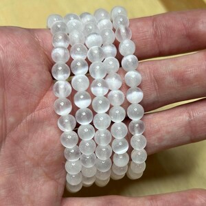 May include: Six white selenite beaded bracelets. Each bracelet is made of round, opaque, white beads. The bracelets are stacked on top of each other. The beads appear to be approximately 0.3 inches in diameter.