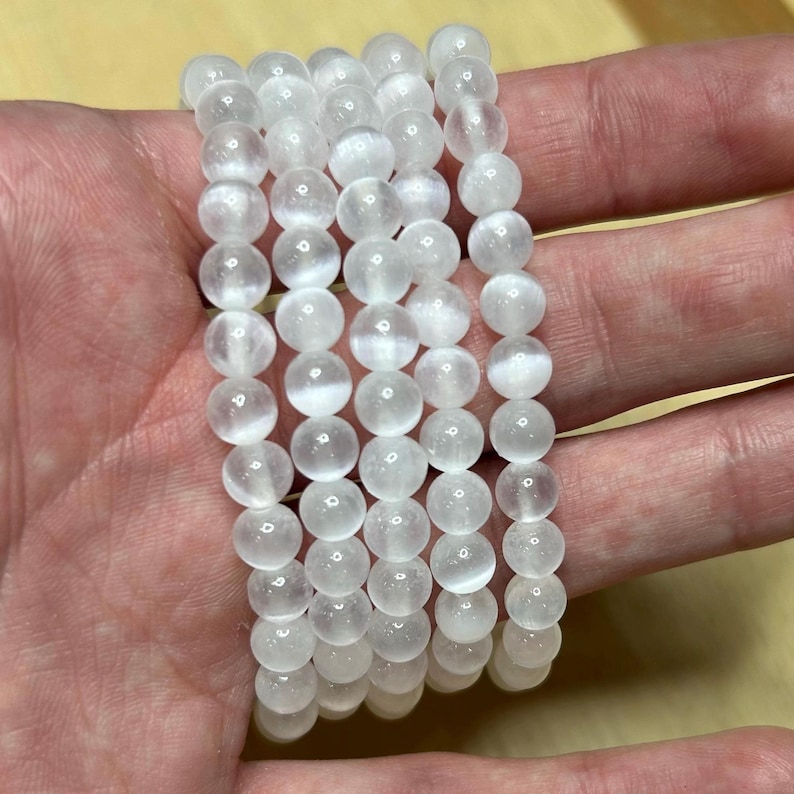 May include: Six white selenite beaded bracelets. Each bracelet is made of round, opaque, white beads. The bracelets are stacked on top of each other. The beads appear to be approximately 0.3 inches in diameter.