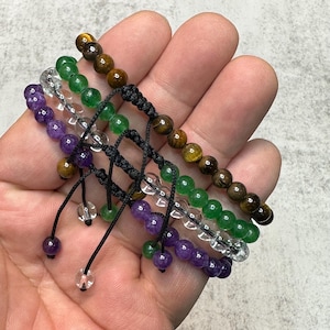 Puede incluir: Una mano muestra cinco pulseras de cuentas. Las pulseras presentan cuentas de varios colores, incluyendo marrón, verde, transparente y morado. Cada pulsera es ajustable con un cordón negro y una cuenta transparente. Las pulseras son una mezcla de piedras naturales y cristales.