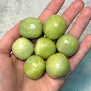 May include: Seven smooth, rounded, light green stones are held in a hand. The stones have a polished appearance and a subtle marbling effect. The overall color is a soft, pale green, reminiscent of jade or a similar gemstone. The stones are a natural, organic shape.