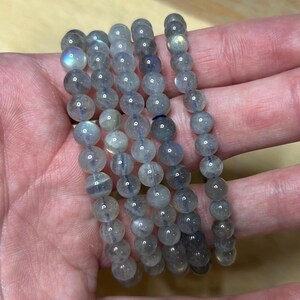 May include: Several strands of round, gray labradorite gemstone beads. The beads have a subtle iridescence, with flashes of blue and green. The beads are strung together, forming bracelets. The bracelets are held in a hand.