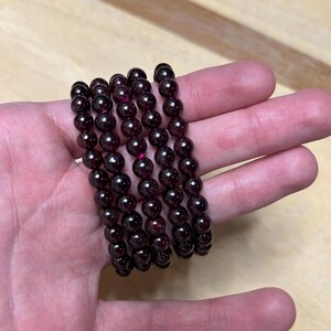 May include: Six strands of dark red garnet gemstone bead bracelets. The beads are round and have a polished finish. The bracelets are held in a hand, showcasing the deep color and texture of the gemstones. The bracelets are a deep, rich red.