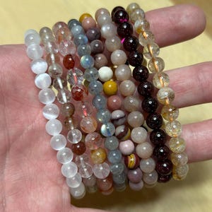 May include: A collection of beaded bracelets in various colors, including white, clear, red, blue, yellow, and brown. The bracelets are made of small, round beads and are held in a hand.