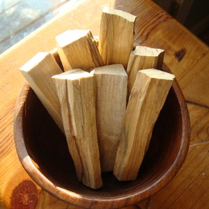 May include: A wooden bowl filled with bundles of dried palo santo sticks. The sticks are light brown and have a natural, rough texture.