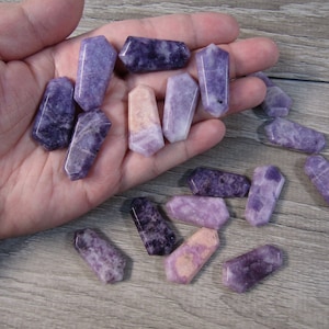 May include: A collection of polished, purple crystals in various shapes and sizes. The crystals are held in a hand, showcasing their varying shades and textures, ranging from deep violet to pale lavender. The crystals are likely used for decorative or metaphysical purposes.
