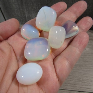 May include: A collection of polished opalite stones in various shapes, including oval and rounded rectangular forms. The stones display a milky white base with iridescent flashes of blue, pink, and yellow. The stones are held in a hand.