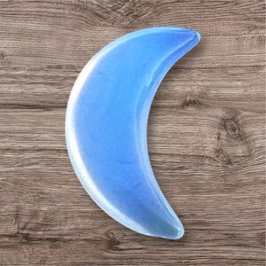 May include: A light blue, crescent-shaped crystal. The polished stone has a smooth, reflective surface and is set against a wooden background. The crystal's shape resembles a crescent moon.