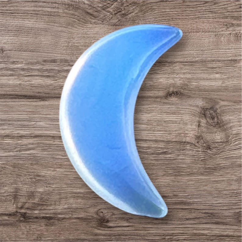 May include: A light blue, crescent-shaped crystal. The polished stone has a smooth, reflective surface and is set against a wooden background. The crystal's shape resembles a crescent moon.
