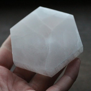 May include: A white, hexagonal crystal, possibly selenite, is held in a hand. The crystal has a smooth, translucent appearance and a geometric shape. The background is a blurred, dark color.