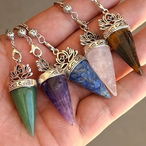 May include: Five cone-shaped crystal pendulums in green, purple, blue, pale pink, and brown tiger's eye. Each stone is capped with a silver-toned metal lotus flower design featuring an Om symbol and attached to a silver-toned chain with a lobster clasp.
