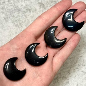 May include: Four polished black obsidian crescent moon shapes. The moons are smooth and reflective, with a dark, glossy finish. The crescent moons are displayed on a light background.