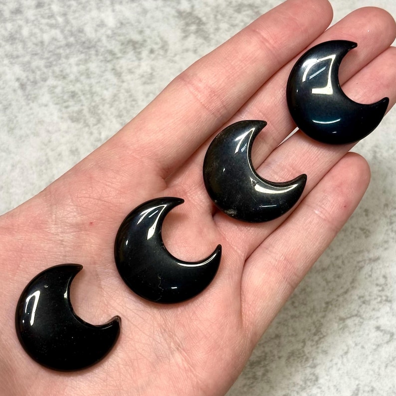May include: Four polished black obsidian crescent moon shapes. The moons are smooth and reflective, with a dark, glossy finish. The crescent moons are displayed on a light background.
