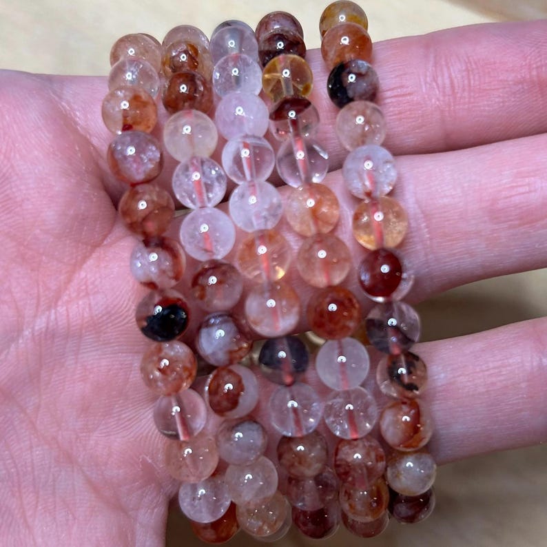 May include: A collection of beaded bracelets featuring translucent, round beads in shades of red, orange, and clear. The beads are strung together, creating a layered effect. The bracelets are displayed on a light-toned surface.