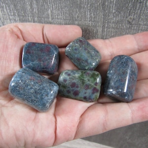 May include: A collection of tumbled stones, each displaying a mix of blue-grey and green hues with red inclusions. The stones are smooth and rounded, held in an open hand, showcasing their natural textures and colors. The stones are approximately 1 inch in length.