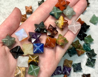May include: A collection of small, star-shaped gemstone carvings in various colors, including green, red, brown, and clear. The gemstones are held in a hand, showcasing their different textures and hues. The carvings are likely used for decorative or spiritual purposes.