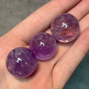 May include: Three polished amethyst spheres, each with a translucent, purple hue. The spheres are held in a hand, showcasing their smooth, rounded forms and internal crystalline structures. The amethyst spheres are a natural gemstone.