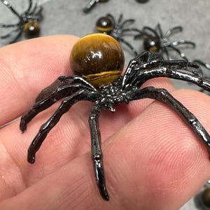 May include: Black metal spider figurine with a round, brown tiger's eye gemstone body. The spider has eight legs and is held in a hand. Other spider figurines are visible in the background. The figurines are likely for Halloween or a similar theme.