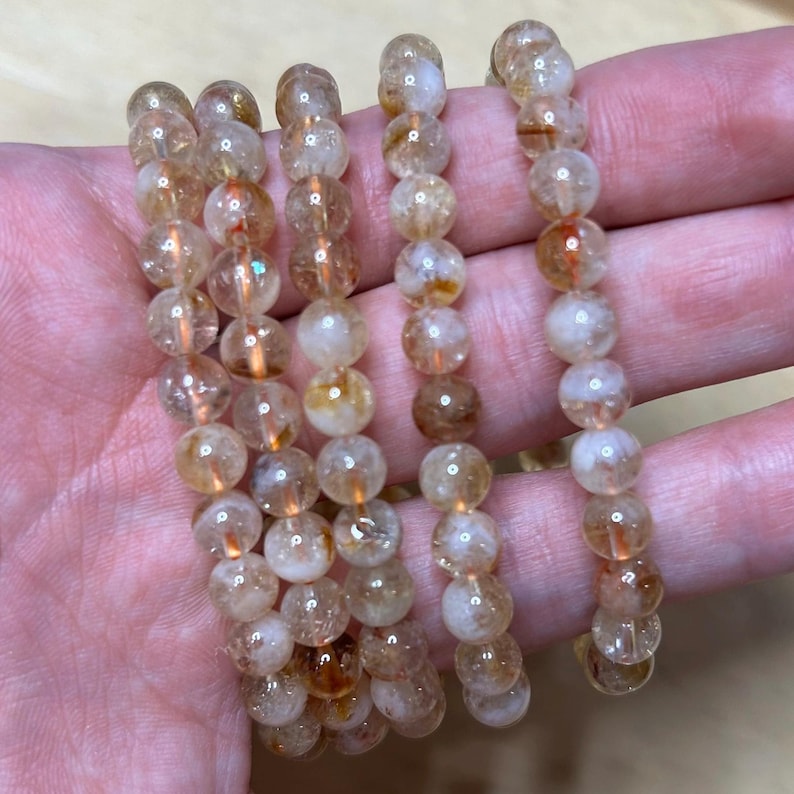 May include: Several strands of beaded bracelets. The beads are a mix of translucent and opaque, with colors ranging from light yellow to amber. The beads are round and appear to be made of a polished stone or crystal material. The bracelets are displayed on a hand.