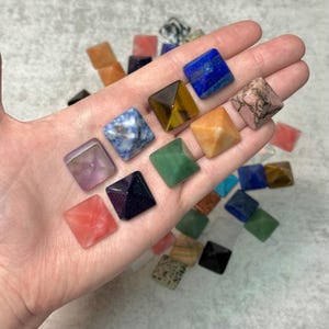 May include: A hand displays a selection of small, square gemstone pyramids in diverse colours. Gemstones include amethyst, blue lapis lazuli, tiger's eye, and rose quartz. Other pyramids are orange, green, and black.