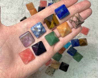 Peut inclure: Une main présente une collection de petites pyramides carrées en pierres précieuses de différentes couleurs. Les pierres comprennent l'améthyste, le lapis-lazuli bleu, l'œil de tigre et le quartz rose. D'autres pyramides sont orange, vertes et noires.