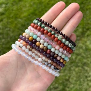 May include: A hand displays a collection of beaded bracelets in various colors. The bracelets feature small, round beads in shades of white, pink, purple, brown, green, and red. The bracelets are likely made of gemstones or other natural materials.