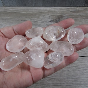 May include: A collection of clear quartz tumbled stones, each with a smooth, rounded shape. The stones vary slightly in size and clarity, with some showing internal inclusions. The stones are held in a hand, showcasing their natural beauty.