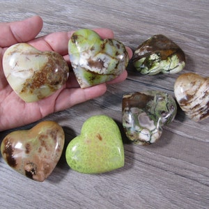 May include: A collection of heart-shaped stones in shades of green, brown, and cream. The stones vary in pattern and texture, with some displaying a marbled appearance. The collection is arranged on a wooden surface, showcasing the natural beauty of the gemstones.