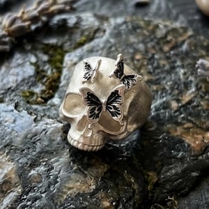 May include: A silver-colored ring in the shape of a skull, adorned with three butterflies. The butterflies have black detailing on their wings. The ring is set against a textured, dark gray background.