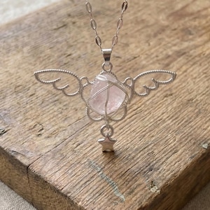May include: A silver-toned necklace featuring a pendant with a pink crystal wrapped in wire, designed with wing-like details. A small star charm hangs below the crystal. The necklace is displayed on a wooden surface.