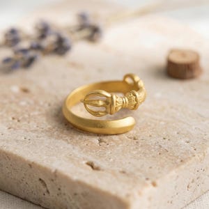 May include: A gold-colored ring with an ornate, detailed design. The ring features a decorative element with a central, stylized shape and intricate patterns. The ring is set against a neutral background, highlighting its metallic sheen and craftsmanship.