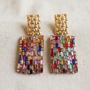 Puede incluir: Un par de pendientes rectangulares dorados. La parte superior tiene un diseño tejido, mientras que la sección inferior está cubierta por un mosaico de gemas rectangulares coloridas. Las gemas incluyen tonos de rojo, azul, verde y rosa.