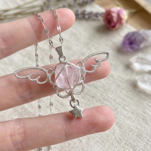 May include: A silver-toned necklace featuring a pink crystal pendant wrapped in wire, with delicate wing-shaped accents. A small star charm dangles below the crystal. The necklace is displayed against a soft, neutral background.