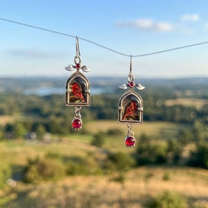 Może przedstawiać: Para srebrnych kolczyków wiszących. Każdy kolczyk przedstawia miniaturowy obraz czerwonego ptaka w dekoracyjnej ramie, z wiszącym czerwonym kamieniem szlachetnym. Kolczyki zawieszone są na rozmytym naturalnym tle.