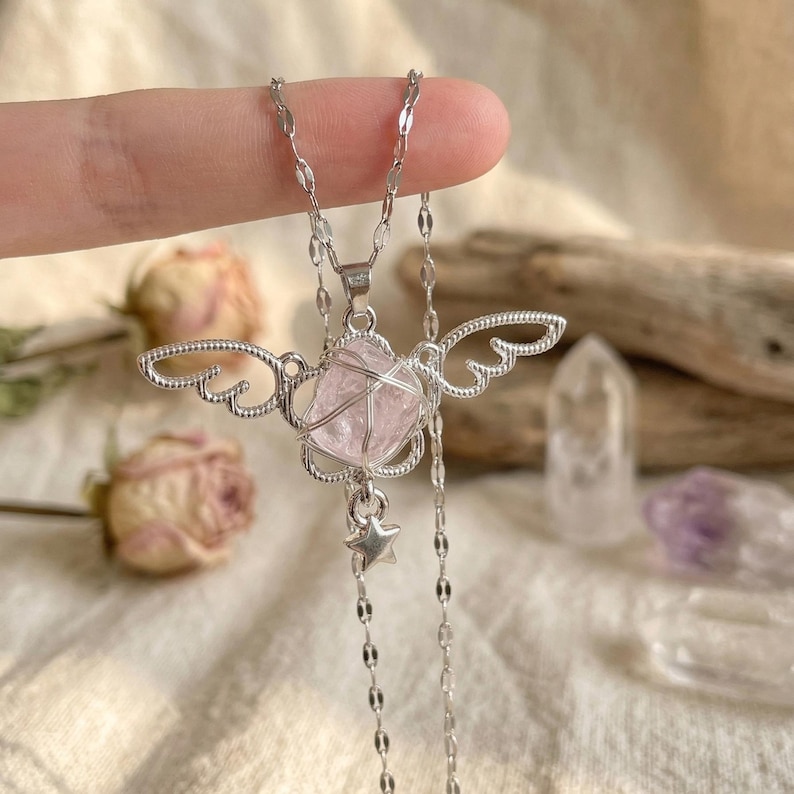 May include: A silver-toned necklace featuring a pink crystal pendant wrapped in wire, with delicate wing-shaped accents and a small star charm. The necklace is displayed against a soft, neutral background, highlighting its intricate design and feminine aesthetic.